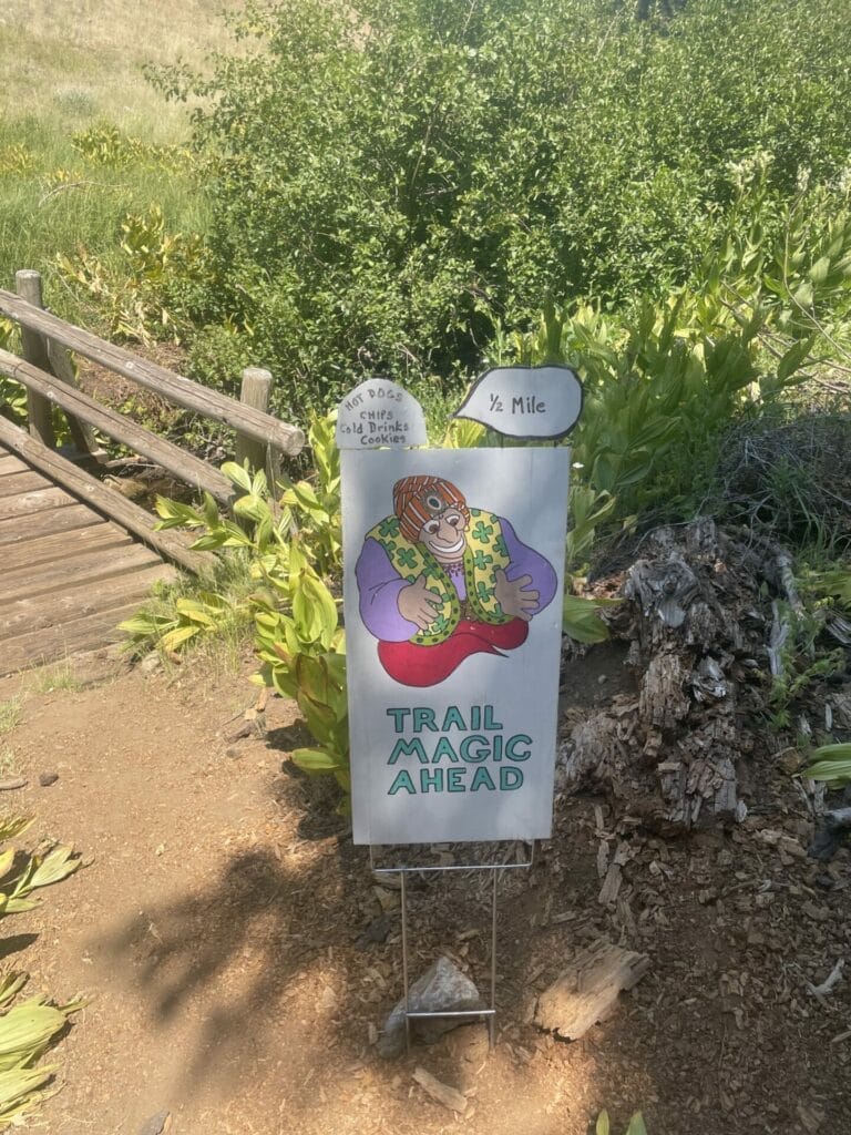 Board sign saying "Trail Magic Ahead" with a gennie drawn on on the trail in front of a foot wooden bridge with greenery around.