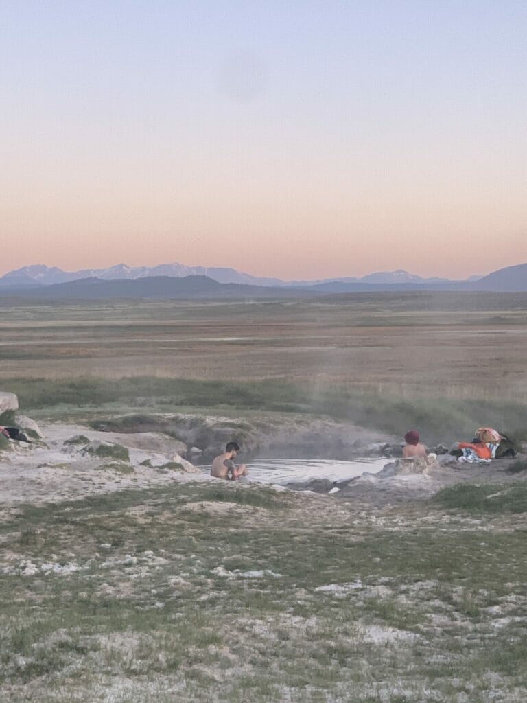 Picture of the hot pool at Wild Willy's Hot Springs with beautiful sunrising sky in the background with a few people in the pool. Here is clothing optional.