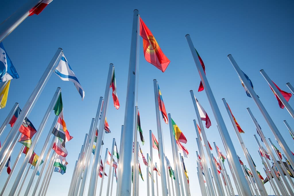 Various countries' flags on flag poles flying in the wind with blue sky in the background.