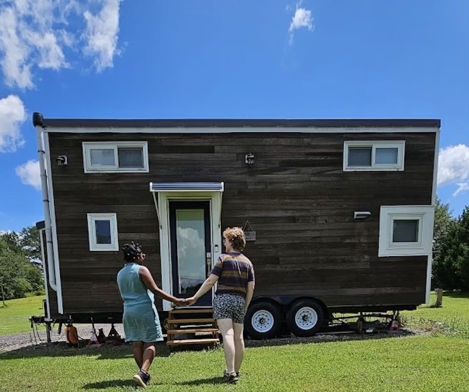 The wooden tiny house with white door and window frames. Author and her spouse holding hands walking towards the front door.