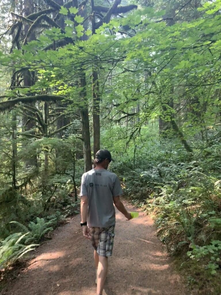 Theron, Lillian's husband, holding a water bottle with a hat on is walking on Rim Trail with lush green heading back to the parking lot.