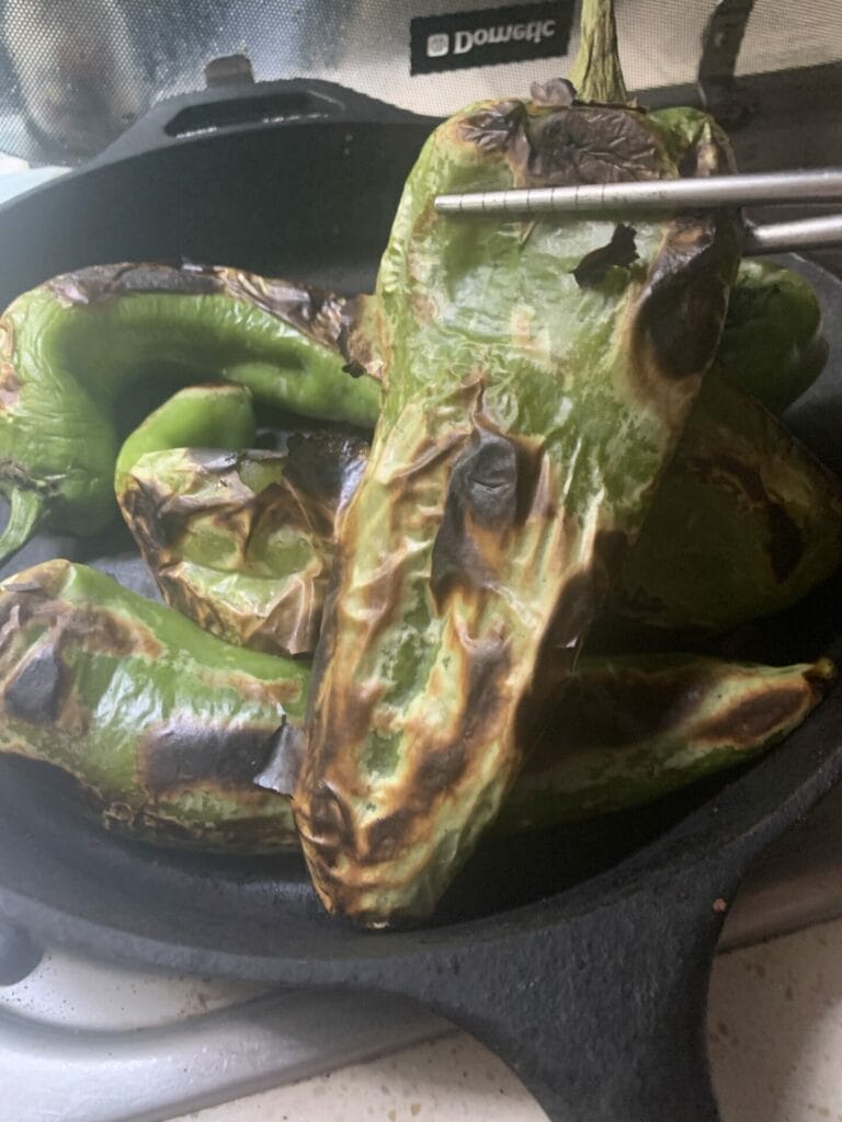 Roasting Hatch chilies in a cast iron pan. One is being held up showing the blisters from the roasting. Yum!