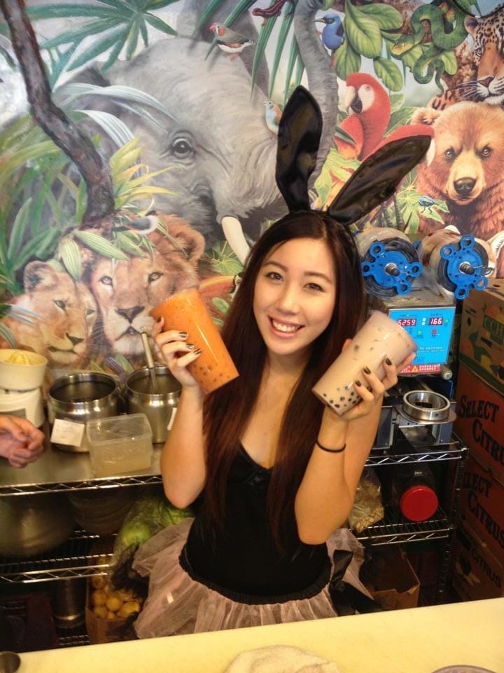 A young woman with bunny ears holding 2 different bubble tea drink with cup sealing machine in the background at a artisan boba shop in Rowland heights, California.